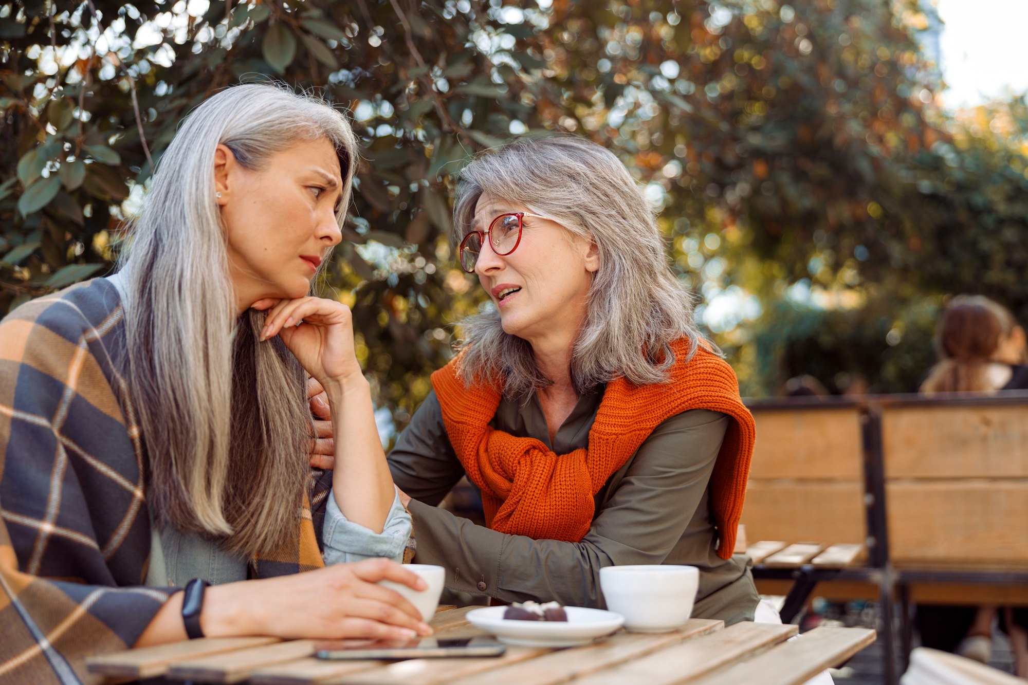 Compasionate senior woman cheers up upset Asian friend sitting together in street cafe